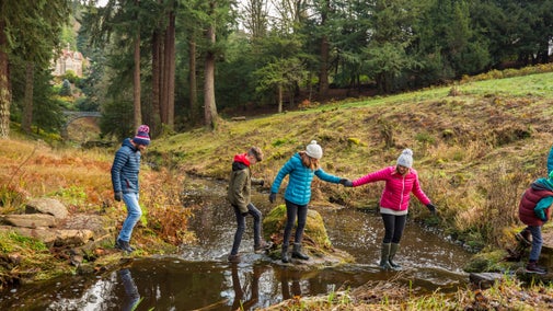 A group of five walkers crossing a small stream in a wooded area, two of them holding hands